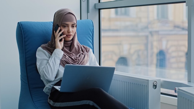 Pensativa gerente árabe feminina em hijab sentada em uma poltrona confortável, conversando por telefone, olhando para o monitor do laptop, considerando uma ideia, uma mulher pensativa tem uma conversa no celular, pensando, tomando uma decisão