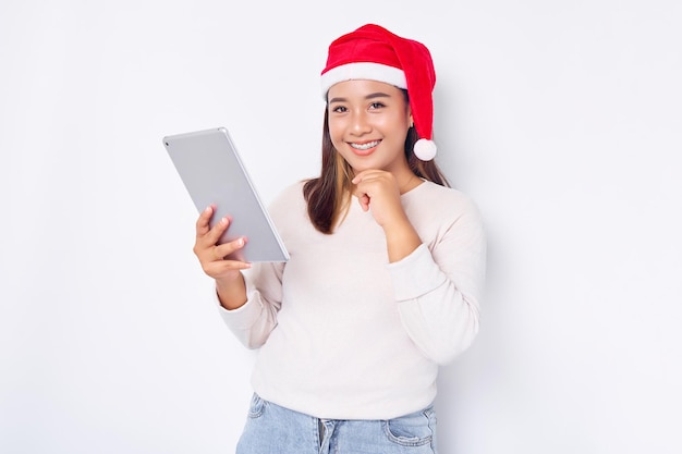 Pensativa jovem asiática em um chapéu de Natal usando um tablet digital e tocando sua bochecha isolada sobre fundo branco Povo indonésio celebra o conceito de Natal