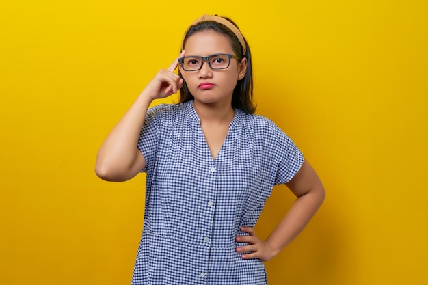 Pensativa jovem asiática usando um vestido xadrez com óculos tentando se lembrar de algo isolado no conceito de estilo de vida de pessoas de fundo amarelo