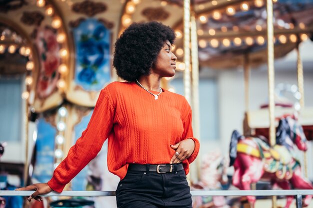 Pensativa jovem negra com penteado afro e suéter vermelho ao lado de um carrossel na rua