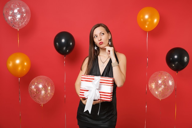 Pensativa jovem num vestido preto comemorando, tentando adivinhar o que está na caixa vermelha com o presente presente nas mãos, em um balão de ar de fundo vermelho brilhante. Feliz Ano Novo, conceito de festa de feriado de maquete de aniversário.