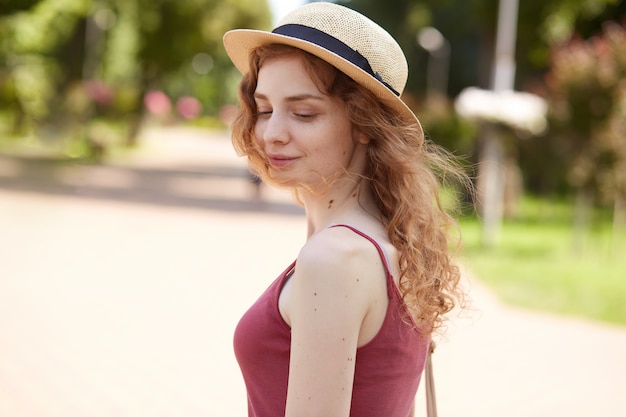 pensativa jovencita atractiva con cabello rubio rizado, posando en el parque, teniendo vacaciones de verano, mirando a un lado