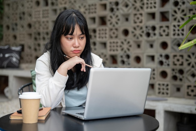 Una pensativa mujer asiática está reflexionando y pensando en su proyecto mientras trabaja en su computadora portátil