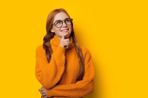 Pensativa senhora caucasiana ruiva está tocando seu queixo enquanto olha pelos óculos e posa em um fundo amarelo com um belo suéter