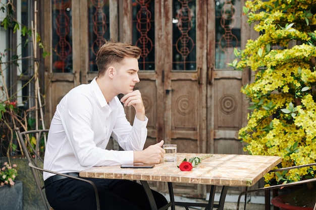 Pensativo apuesto joven sentado a la mesa en un café al aire libre y mirando la rosa roja de su novia