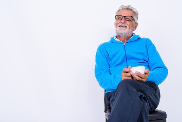 Foto pensativo feliz sênior barbudo homem sorrindo, segurando a xícara de café e sentado na cadeira, pronto para a ginástica em branco