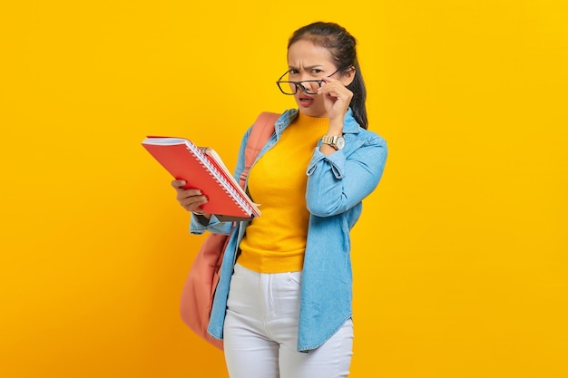 Pensativo hermosa joven estudiante asiática en ropa de mezclilla con mochila sosteniendo cuaderno y mirando a la cámara aislada sobre fondo amarillo Educación en concepto de colegio universitario de secundaria