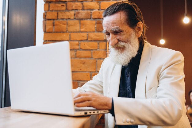Pensativo hombre senior, con una barba blanca, sonríe mirando su portátil. Una persona mayor de raza blanca. Persona mayor y nueva tecnología.