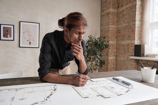 Foto pensativo jovem arquiteto de raça mista com cabelos tingidos, inclinando-se sobre a mesa e analisando o esboço enquanto pensava em design