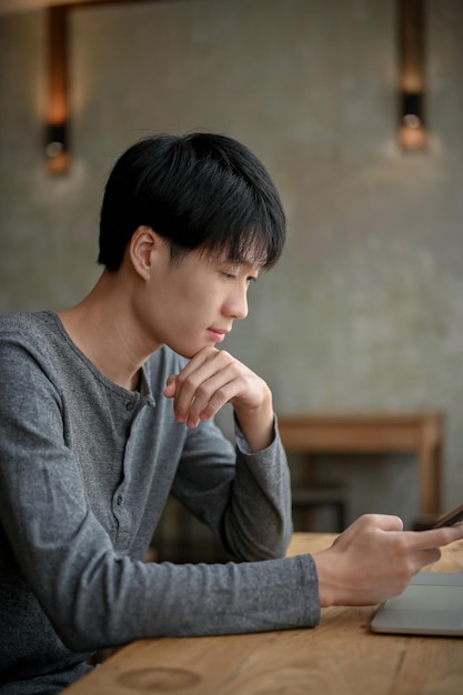 Pensativo jovem asiático pensando pensativamente em algo enquanto olha para a tela do smartphone