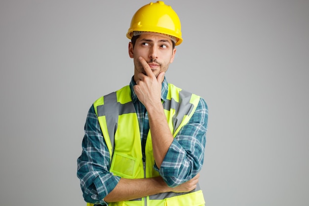 Pensativo jovem engenheiro masculino usando capacete de segurança e uniforme mantendo a mão no queixo olhando para cima isolado no fundo branco