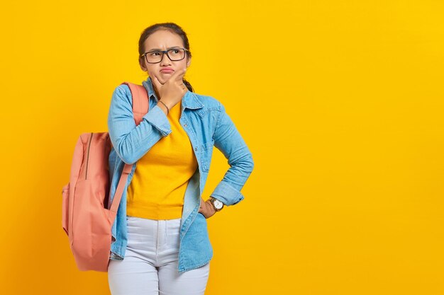 Pensativo jovem estudante asiático em roupa jeans com mochila segurando o queixo enquanto pensava em algo isolado em fundo amarelo Educação no conceito de faculdade universitária do ensino médio