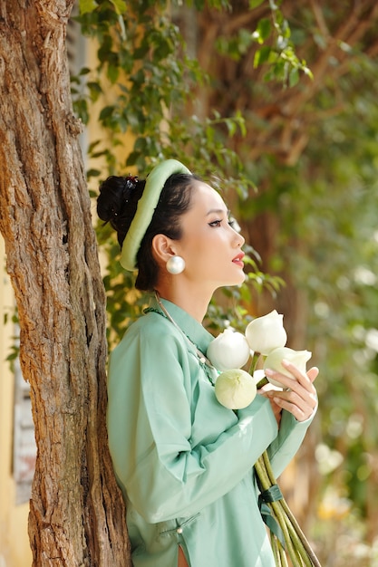 Pensativo joven y bella mujer vietnamita en traje tradicional sosteniendo ramo de flores y apoyado en el árbol