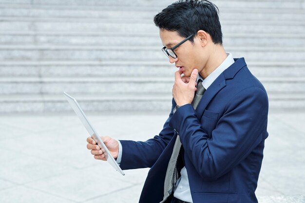Pensativo joven empresario vietnamita en gafas frotando la barbilla y leyendo el correo electrónico en la tableta