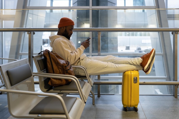 Pensativo joven hipster negro sentado en la terminal del aeropuerto usando un teléfono inteligente y esperando aterrizar
