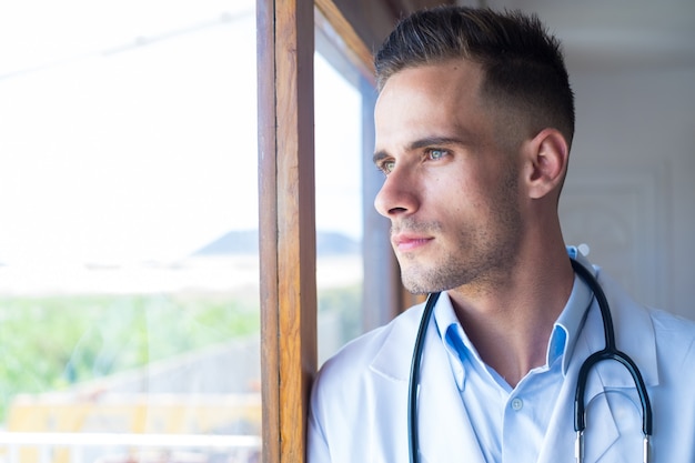 Pensativo joven médico caucásico en uniforme médico blanco mira en la ventana a distancia pensando o reflexionando, hombre serio GP planifica la carrera futura o el éxito en la medicina, visualiza en la escritura del lugar de trabajo