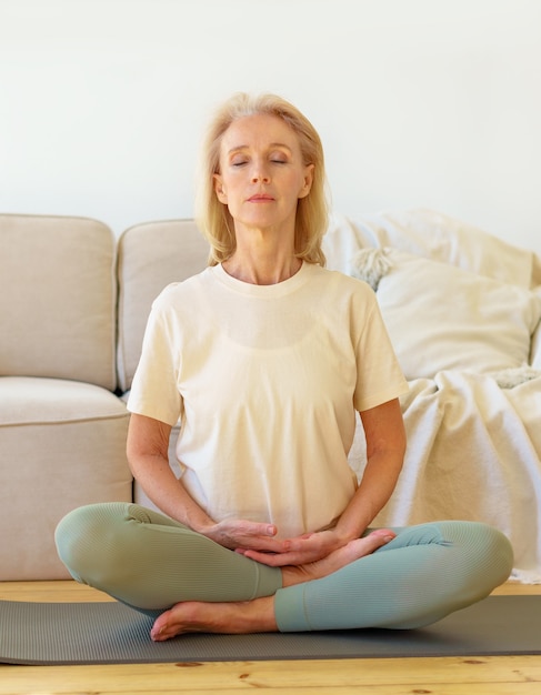 Pensionierte Frau, die Yoga meditiert und praktiziert, während sie in der Lotuspose auf dem Boden zu Hause vertikal sitzt sitting