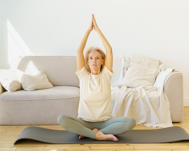 Pensionierte Frau im Lotussitz praktiziert Yoga-Übungen zu Hause auf Matte sitzend