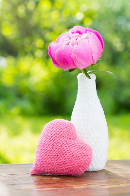 Peonía rosa en un jarrón blanco decorado con tejido de punto y un corazón de punto rosa sobre una mesa de madera