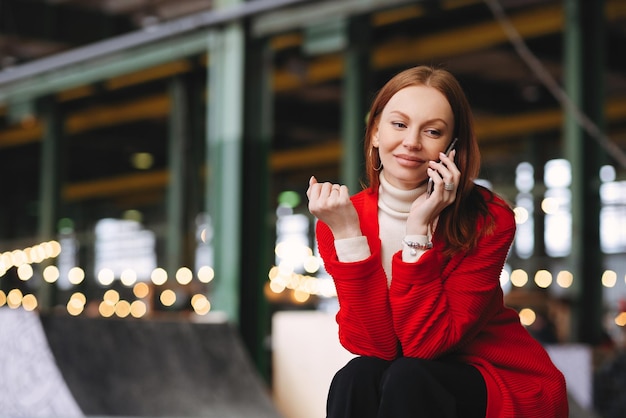 People Technology und Lifestyle-Konzept Eine positive kaukasische Frau führt Telefongespräche mit Freunden über das Mobiltelefon und trägt einen roten Mantel und eine schwarze Hose, die draußen mit freiem Platz posiert