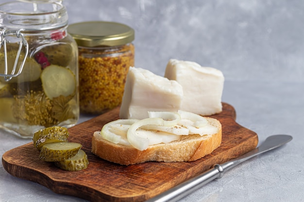 Pepinos encurtidos en un frasco de vidrio sobre una tabla de madera y un sándwich con tocino. Habas de mostaza en el fondo. Comida fermentada. Copie el espacio.