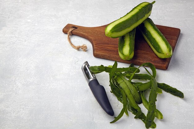 Pepinos frescos con piel y cuchillo sobre una tabla de cortar de madera marrón sobre una mesa de hormigón gris claro