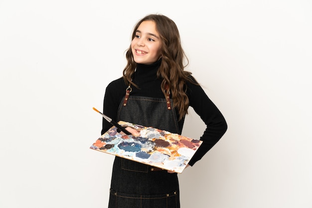 Pequeña artista mujer sosteniendo una paleta aislada en la pared blanca posando con los brazos en la cadera y sonriendo