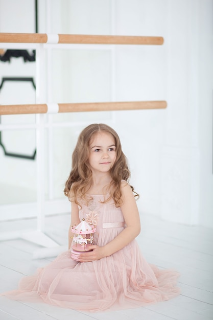 Una pequeña bailarina con un vestido se sienta en una clase de baile en el suelo. Niña sosteniendo un carrusel de juguete musical. El niño recibe un regalo. Juguete de carrusel musical vintage. sala de clase de salón de ballet interior