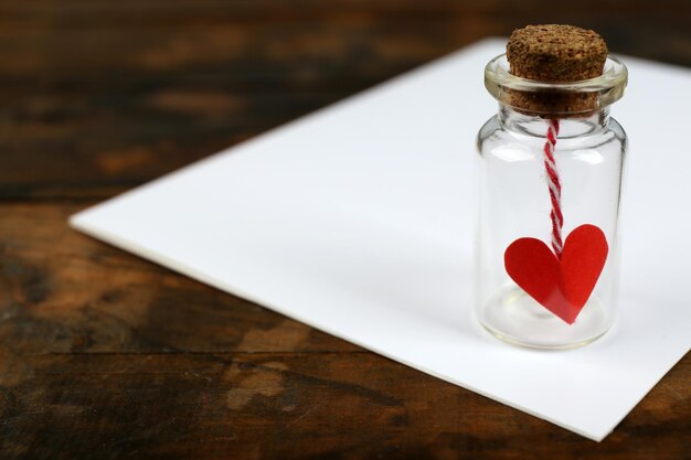 Pequeña botella de vidrio con corazón en una hoja de papel en blanco sobre fondo de mesa de madera rústica