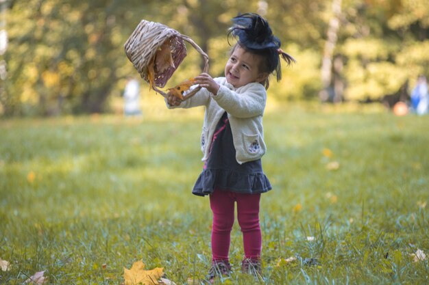 pequeña bruja linda jugando con una canasta de hojas en el parque de otoño.