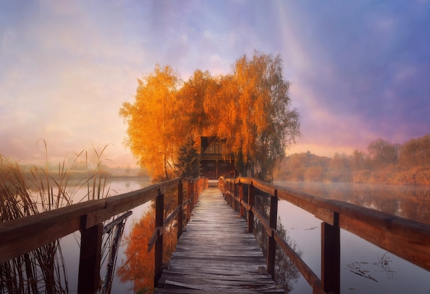 Pequeña casa de madera en la isla con un muelle de madera en medio de un lago en la brumosa mañana de otoño durante el amanecer. Paisaje de hadas en otoño.