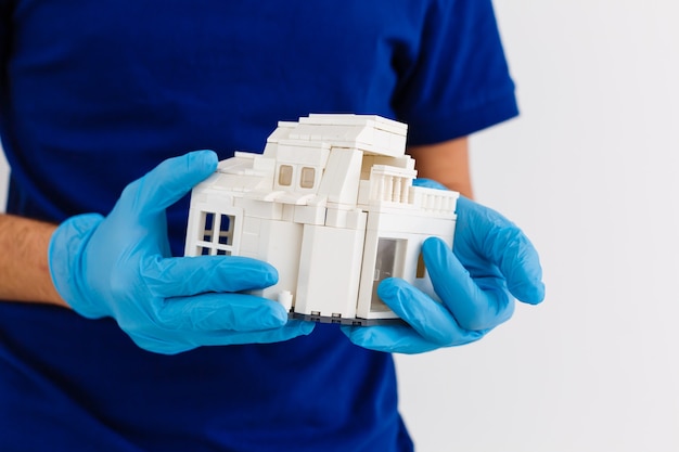 pequeña casa de madera en la palma de su mano con un guante de goma médico blanco, fondo azul, modelo de madera de la casa, concepto de cuarentena, pandemia de coronavirus, medicina durante una epidemia