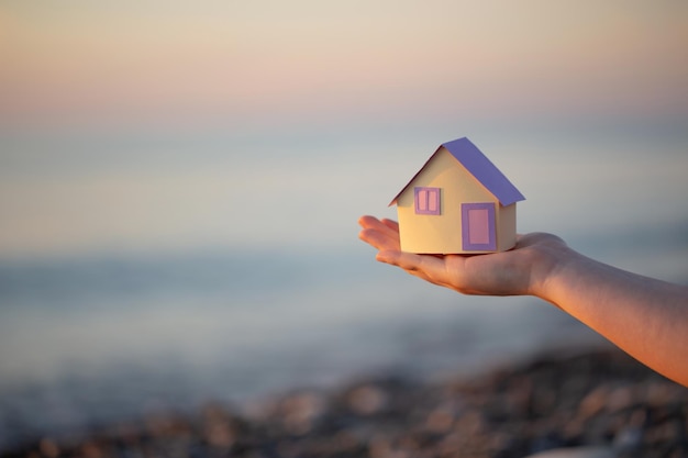 Una pequeña casa de papel en la mano contra el fondo del mar al atardecer el concepto de servicios inmobiliarios un sueño de una casa junto al mar