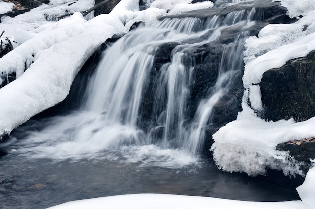 Una pequeña cascada de montaña está cubierta de nieve. Arroyo en el bosque, paisaje de invierno, fondo claro