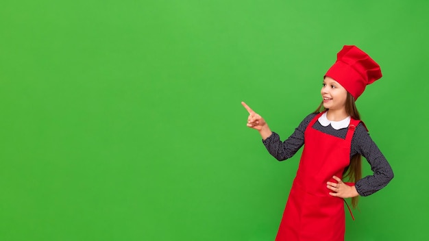 Una pequeña chef con un delantal y una gorra de cocina apunta a su anuncio en un fondo verde aislado Copiar espacio