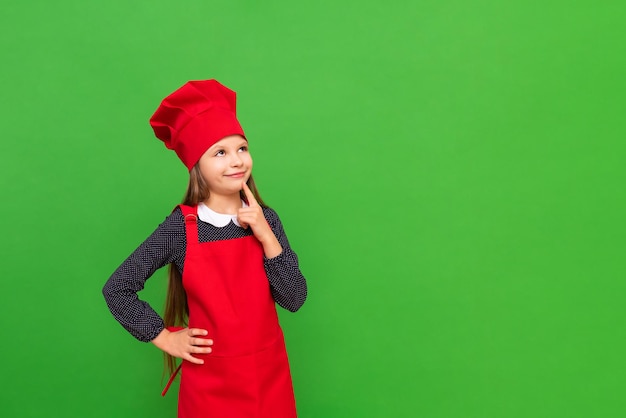 Una pequeña chef con un delantal y una gorra de cocina apunta a su anuncio en un fondo verde aislado Copiar espacio