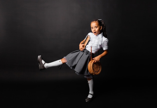 Pequeña colegiala alegre con libro y bolso volviendo a la escuela en la nueva temporada aislado sobre fondo oscuro