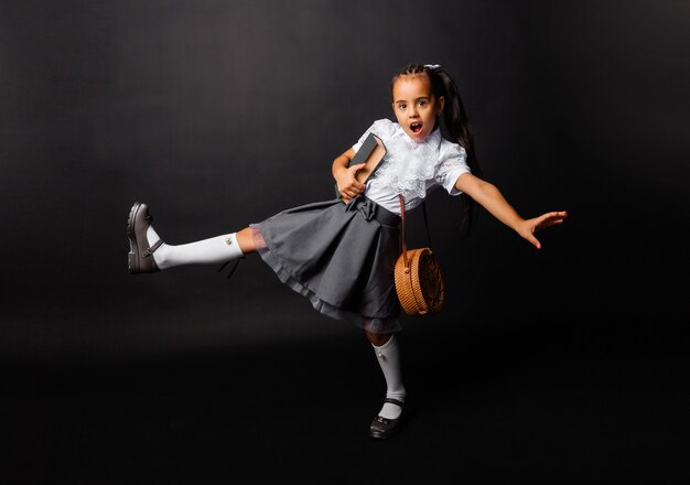 Pequeña colegiala alegre con libro y bolso volviendo a la escuela en la nueva temporada aislado sobre fondo oscuro