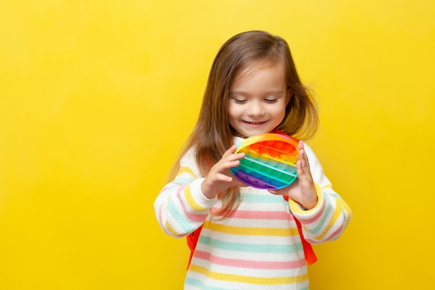 Una pequeña colegiala con una blusa a rayas y con una mochila sostiene un pop en sus manos y sonríe