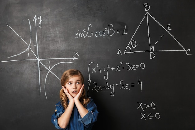 Pequeña colegiala inteligente de pie en la pizarra con gráficos matemáticos escritos en él