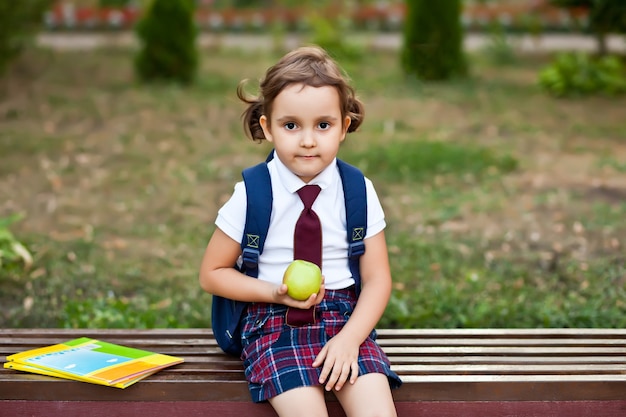 Pequeña colegiala linda en uniforme sentado en un banco y comiendo una manzana