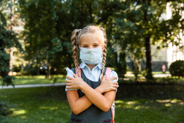 Pequeña colegiala con una máscara médica se sienta en el parque sobre el césped y se divierte.