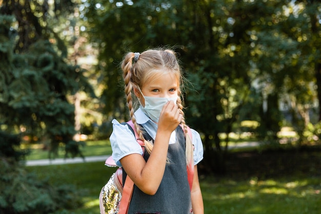 Pequeña colegiala con una máscara médica se sienta en el parque sobre el césped y se divierte.