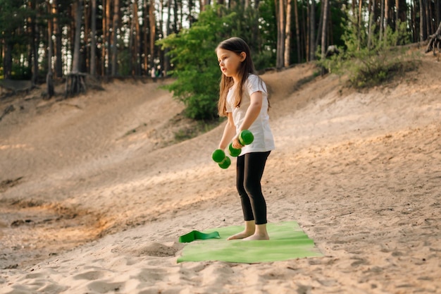 Foto pequeña dama trabajando con pesas en las manos de pie en una alfombra deportiva al aire libre entrenamiento de atleta junior en una playa de arena