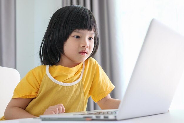 Una pequeña estudiante asiática estudia en línea usando una computadora portátil en casa