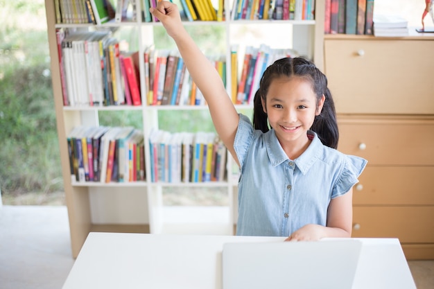 Pequeña estudiante con la computadora portátil en el escritorio en la biblioteca y fighto actuando