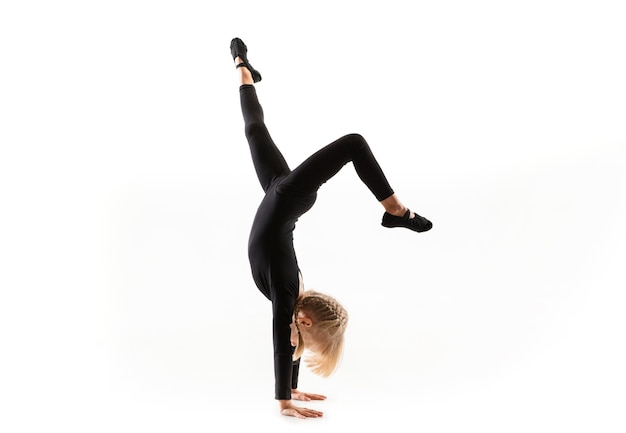 Pequeña gimnasta flexible femenina está practicando aislado sobre fondo blanco. Chica rubia caucásica en entrenamiento deportivo negro en ejercicios de atletismo. Concepto de deporte, infancia, estilo de vida saludable.