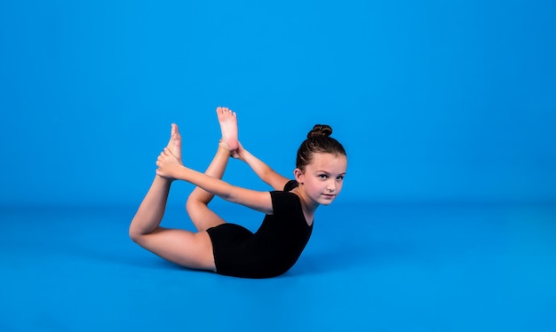 Una pequeña gimnasta en traje de baño negro realiza ejercicios de flexibilidad sobre un fondo azul con un lugar para el texto