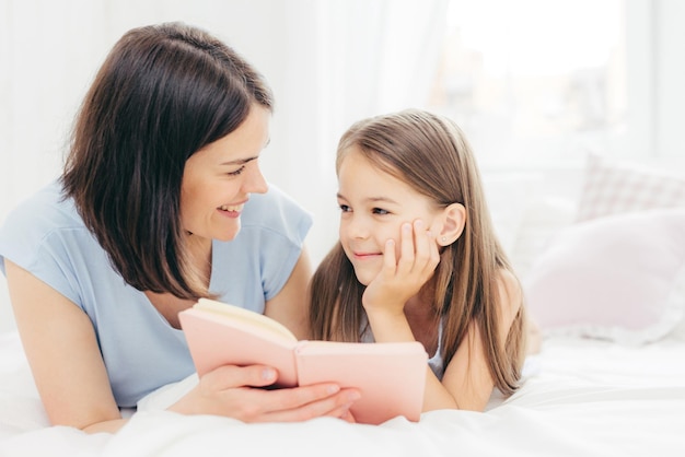 La pequeña hija complacida mira con curiosidad a su madre que lee cuentos de hadas sostiene un pequeño libro en una cama cómoda disfruta de la unión Un niño pequeño y alegre se prepara para la escuela Concepto de paternidad