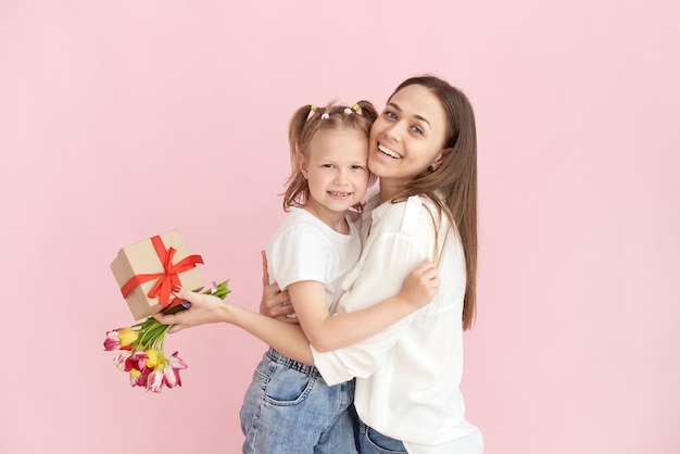 Una pequeña hija le da flores a su madre y un regalo para el día de la madre sobre un fondo rosa en el estudio Retrato feliz de mamá e hija